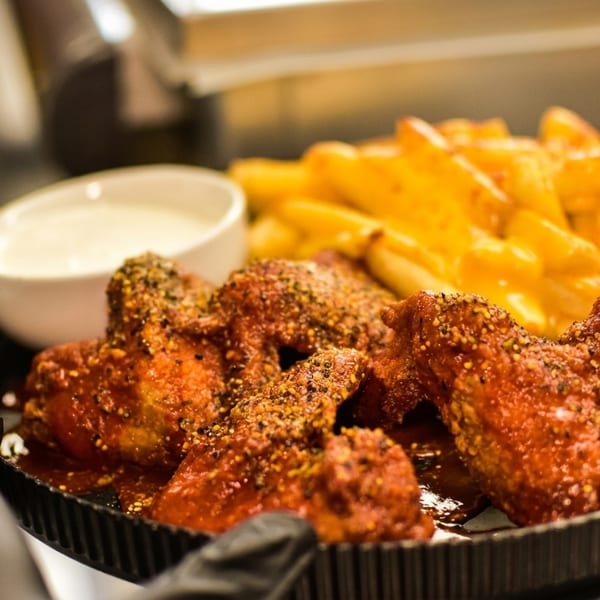 A gloved hand holds a plate of spicy chicken wings, cheese fries, and a side of ranch.